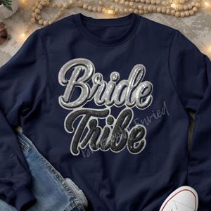 May include: A navy blue sweatshirt with the words "Bride Tribe" in a silver and black glitter design. The sweatshirt is folded with a pair of blue jeans and white sneakers.