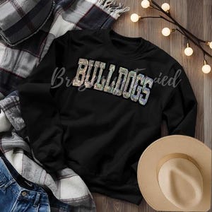 May include: Black crewneck sweatshirt with the word "BULLDOGS" in a shimmering, iridescent font. The sweatshirt is displayed with a plaid blanket, a pair of jeans, and a tan hat. The word "Bright" is written in cursive on the top left of the sweatshirt.