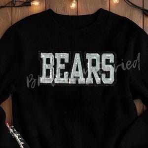 May include: Black crew neck sweatshirt with the word "BEARS" in a silver, textured, block letter design across the chest. The sweatshirt is made of a soft, ribbed material.