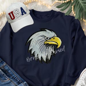 May include: Navy blue sweatshirt featuring an embroidered eagle head with gray and yellow details. A white baseball cap with "USA" in red and blue lettering is next to the sweatshirt. The text "Braved" is written in cursive.