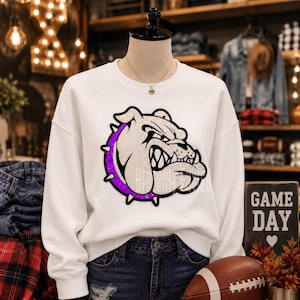 May include: A white long-sleeve sweatshirt featuring a large graphic of a bulldog head in profile. The bulldog has a black outline and a purple spiked collar. The design is rendered in a textured, shimmering material on the front of the garment.