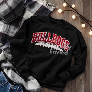 May include: A black long-sleeve sweatshirt featuring the word BULLDOGS in red, textured, varsity-style lettering arched over a white graphic of a football. The design is centered on the chest of the garment, which has a crew neckline.