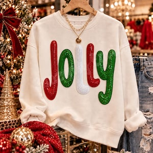 May include: A cream-colored crewneck sweatshirt featuring the word JOLLY in large, colorful, sequined letters. The J is red, the O is green, the L is white, the L is red, and the Y is green. A gold chain necklace rests at the neckline.