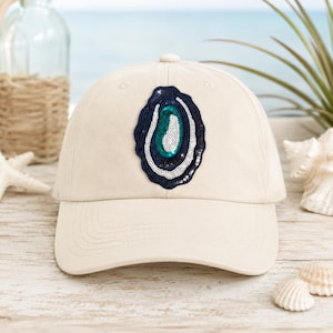May include: A cream-colored baseball cap featuring a central embroidered patch shaped like an oyster shell. The patch is decorated with concentric layers of dark navy blue, white, and teal green sequins, creating a textured, shimmering appearance.