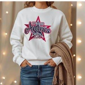 May include: White sweatshirt with a red and blue star graphic. The graphic features the word "America" in a script font, along with the numbers "1776-2026" and "250". A brown scarf is draped over the arm.
