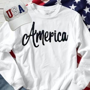 May include: A white sweatshirt with the word "America" in black script. A white baseball cap with "USA" in red and blue lettering is to the left. The background includes a portion of an American flag.