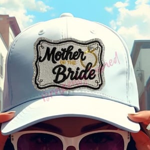 May include: A light blue baseball cap with the text "Mother of the Bride" in black script, framed by a silver glitter design and a gold ring. The cap is a classic style, perfect for a wedding.
