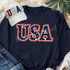 May include: Navy blue sweatshirt with a sequined "USA" design in red, white, and blue. A white baseball cap with "USA" embroidered in red and blue is next to the sweatshirt. The image also includes blue jeans and white sneakers.