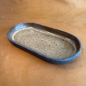 May include: A handmade, oval ceramic tray. The interior is speckled light brown, and the exterior is dark brown and textured. The rim is a darker shade of brown. The tray is displayed on a brown surface.