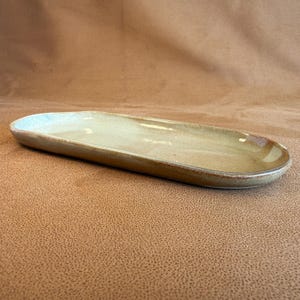 May include: A handmade, oblong ceramic serving dish. The dish has a light tan interior with a darker brown rim. The ceramic dish is a neutral colour and would be suitable for serving appetizers or snacks.