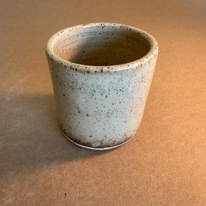May include: A handmade ceramic tumbler with a speckled, off-white glaze. The cylindrical cup has a slightly tapered shape and a white base. The rim is a darker brown colour. This rustic mug is perfect for coffee, tea, or other beverages.
