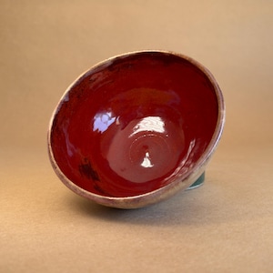 May include: A handmade ceramic bowl with a glossy, deep red interior and a textured, bronze-coloured rim. The bowl is tilted, revealing its interior. The exterior has a green base. This unique bowl is perfect for serving or display.