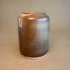 May include: A cylindrical ceramic vase with a speckled brown and tan glaze. The vase has a wide opening and a white base. The glaze has a drip effect on one side. The vase is approximately 18 cm tall.