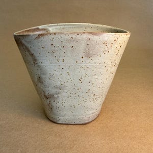 May include: A speckled, off-white ceramic vase with a wide, slightly irregular opening. The vase has a conical shape with horizontal grooves and brown speckles. The interior is a darker shade of the same off-white colour.