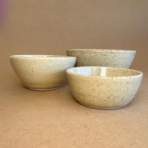 May include: Three speckled ceramic bowls in varying sizes. The bowls are a light beige colour with small brown speckles. The bowls are arranged in a group, with the largest bowl at the back.