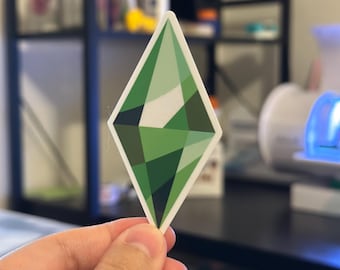 May include: A diamond-shaped sticker featuring a geometric design in shades of green and white. The sticker has a white border and is held in a hand. The design is reminiscent of a video game icon.