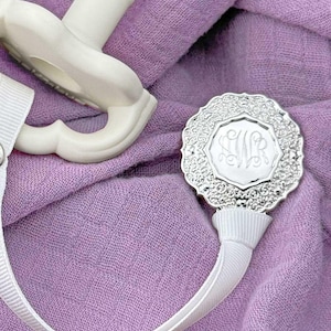 May include: A white baby pacifier with a matching ribbon and a silver-colored clip. The clip has an ornate, floral-like border and a monogrammed center. The pacifier and clip are on a lavender fabric background.