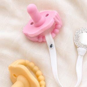 May include: A close-up of three baby pacifiers in pink and yellow. The pink pacifier has a matching clip and a beaded edge. The yellow pacifier has a beaded edge. A silver clip is also visible.