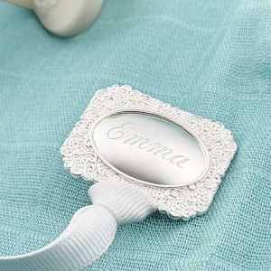 Puede incluir: Una etiqueta plateada y ornamentada con el nombre "Emma" grabado en un centro ovalado. La etiqueta está sujeta a una cinta blanca y descansa sobre una tela azul claro. La etiqueta tiene un borde decorativo, similar a una flor.