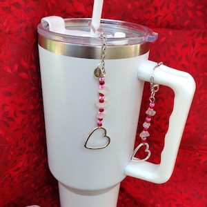 May include: White stainless steel tumbler with a lid and a handle.  Two silver charms with pink and white beads hang from the lid and handle.  The charms feature a heart shape at the bottom.
