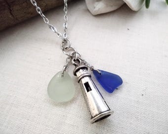 May include: A silver-toned metal necklace featuring a lighthouse charm suspended from a delicate chain. The pendant is accented by two small, smooth pieces of sea glass, one in a pale frosted white and the other in a vibrant, deep cobalt blue.