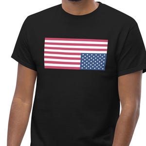 Puede incluir: Camiseta negra con un diseño rectangular de la bandera estadounidense. La bandera tiene rayas rojas y blancas y un cuadrado azul con estrellas blancas. La camiseta está hecha de un material suave.