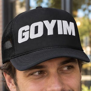 May include: A black trucker hat featuring a mesh back and a solid front panel with the word GOYIM printed in bold white capital letters. The cap has a curved brim and a structured crown, providing a classic look with breathable mesh side panels.