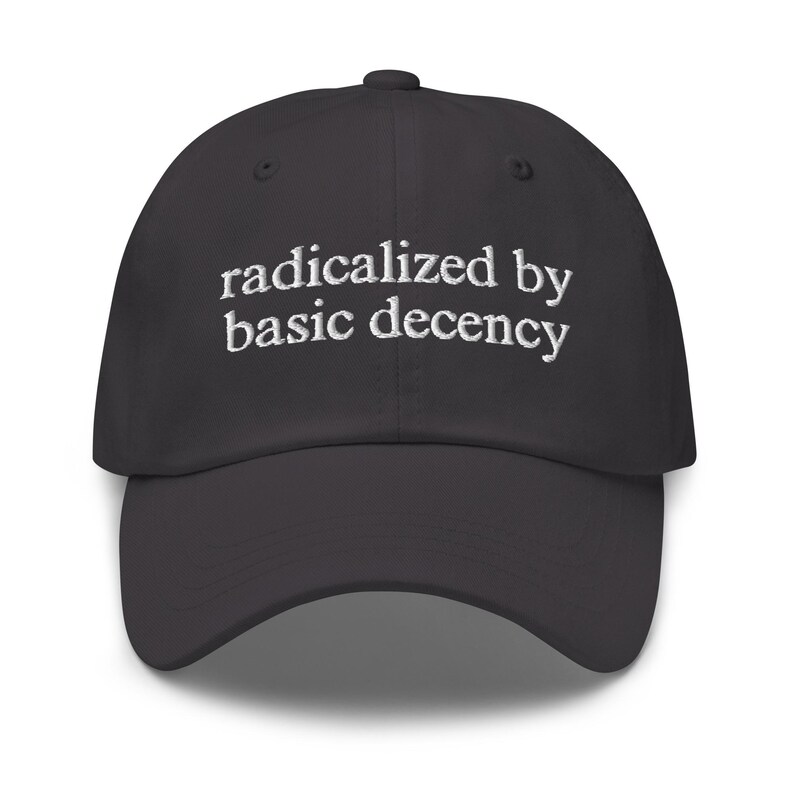 May include: A dark grey baseball cap featuring the embroidered phrase "radicalized by basic decency" in white. The cap has a curved peak and a button on top. A fashionable accessory for casual wear.