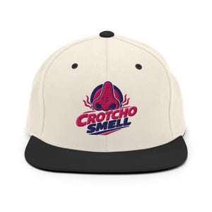 Può includere: Cappellino da baseball a tesa piatta color crema con visiera nera. Il cappellino presenta una grafica di un naso rosso con la scritta "Crotcho Smell" in rosa e blu.