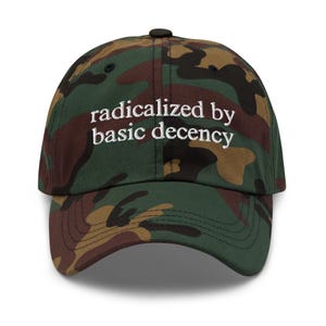 May include: A camouflage baseball cap with the text "radicalized by basic decency" embroidered in white. The cap has a curved peak and a black button. The hat is a mix of green, brown, and tan.