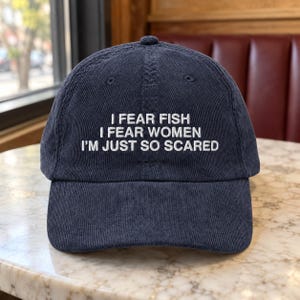 May include: A navy blue corduroy baseball cap featuring white embroidered text that reads: I FEAR FISH, I FEAR WOMEN, I'M JUST SO SCARED. The hat has a structured crown, a curved brim, and a button on top, showcasing a textured fabric finish.