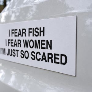 Puede incluir: Una pegatina rectangular blanca con texto negro que dice: I FEAR FISH, I FEAR WOMEN, I'M JUST SO SCARED. La pegatina está aplicada sobre una superficie blanca brillante, mostrando un diseño limpio y contrastado con letras negritas.