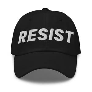 K&ouml;nnte beinhalten: Schwarze Baseballkappe mit dem Wort "RESIST" in Wei&szlig; auf der Vorderseite gestickt. Die Kappe hat einen gebogenen Schirm und einen Knopf oben. Ein schlichtes, k&uuml;hnes Design.