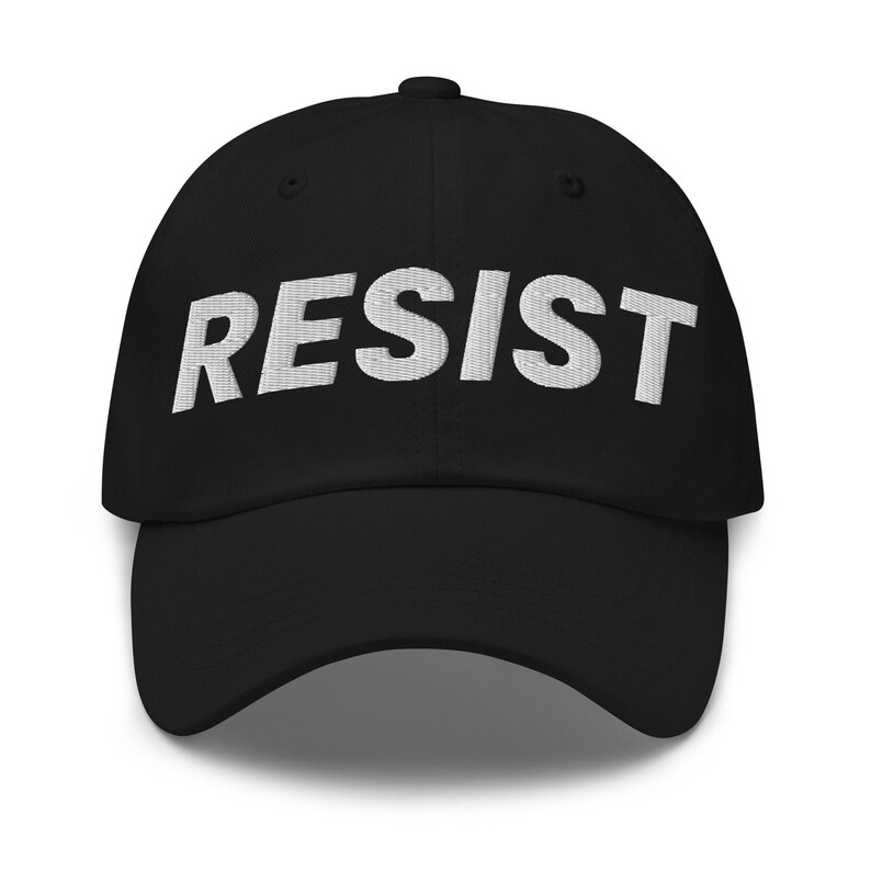 K&ouml;nnte beinhalten: Schwarze Baseballkappe mit dem Wort "RESIST" in Wei&szlig; auf der Vorderseite gestickt. Die Kappe hat einen gebogenen Schirm und einen Knopf oben. Ein schlichtes, k&uuml;hnes Design.