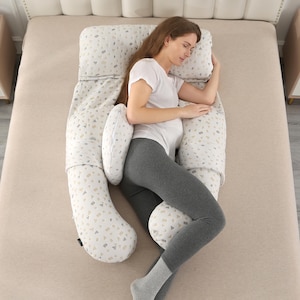 May include: A beige and white patterned pregnancy pillow in a U-shape, designed for side sleeping. The pillow is supporting a person lying on a bed, wearing a white t-shirt and gray leggings. The pillow has a geometric pattern.