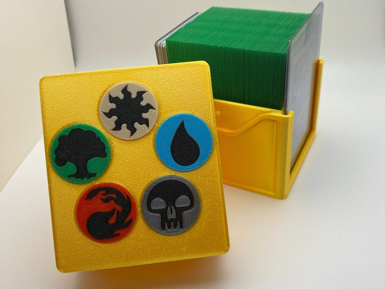 May include: A yellow card game box with five different colored circles containing symbols. The circles are green, white, blue, red, and gray. The symbols are a tree, a sun, a water drop, a flame, and a skull. The box is open and a green deck of cards is visible inside.