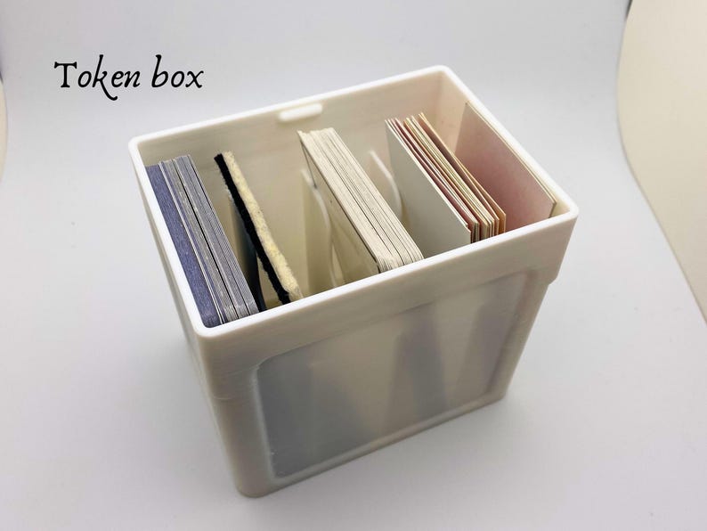 May include: White plastic box with the text "Token box" printed on the front. The box is filled with stacks of cards.
