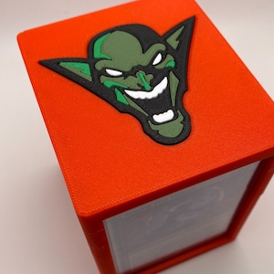 May include: A vibrant orange card storage box featuring a green goblin face design. The goblin face has black, green, and white details. The box is likely plastic and is designed to hold cards, possibly for a game.