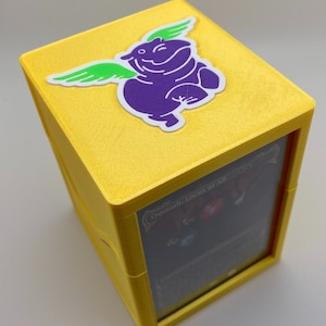 May include: Yellow card game box with a purple hippo with green wings sticker on the top. The box has a clear window on the side showing a card with the text "Omniatic Locus of All".