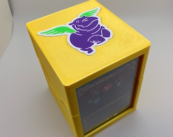 May include: Yellow card game box with a purple hippo with green wings sticker on the top. The box has a clear window on the side showing a card with the text "Omniatic Locus of All".