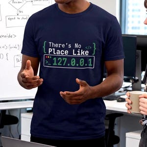 May include: A navy blue short-sleeved t-shirt featuring white and green text that reads "There's No Place Like 127.0.0.1" with curly brackets, a command prompt symbol, and code tags printed on the front of the chest area.