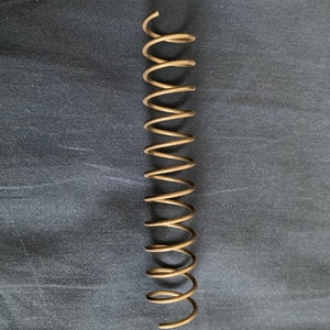 May include: A bronze-colored coil spring, vertically oriented against a dark blue background. The spring has a spiral design with evenly spaced coils. The metal has a slightly aged appearance.