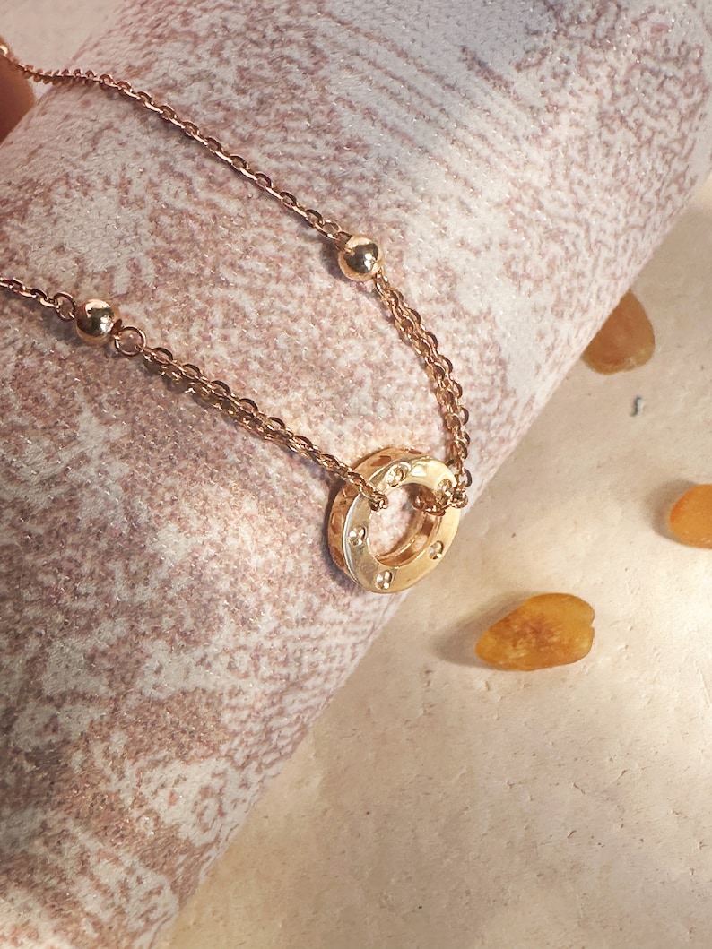 May include: A gold chain necklace with a circular pendant featuring a repeating pattern of small circles. The pendant is hanging on a textured beige surface.