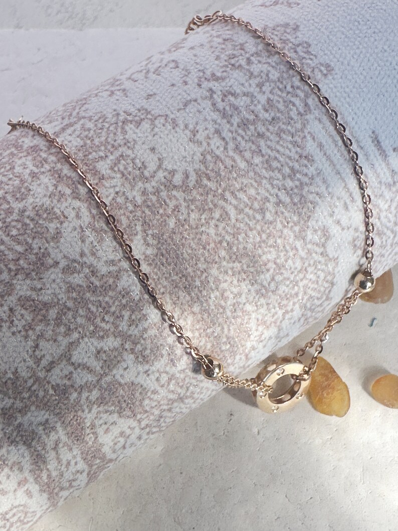 May include: A gold chain necklace with a circular pendant featuring a circle of small diamonds. The necklace is on a white and brown textured surface.