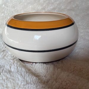 May include: A white ceramic planter with a wide, round shape. The planter has a thick orange band around the top and two black bands around the middle.