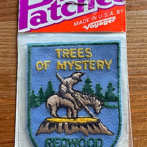 May include: Embroidered patch with a blue background featuring the text "TREES OF MYSTERY" and "REDWOOD HWY." A figure on horseback is depicted in the center, with trees in the background. The patch is in a clear plastic package with an orange header.