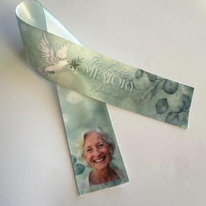 May include: A light green satin ribbon featuring a printed white dove carrying an olive branch, a photograph of a smiling person with white hair, and the text In Loving MEMORY Name, decorated with a subtle green eucalyptus leaf pattern.