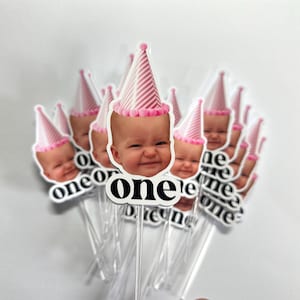 May include: A collection of cake toppers featuring a photographic cutout of a baby's face wearing a pink and white striped party hat with a pink pom-pom trim. Below the face, the word one is printed in a bold, black, lowercase sans-serif font.