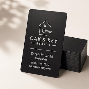 May include: A stack of matte black business cards with rounded corners. The top card features a white line-art logo of a house with a key, the text OAK & KEY REALTY, and the contact details for Sarah Mitchell, Real Estate, (555) 214-7830, and oakandkeyrealty.com.