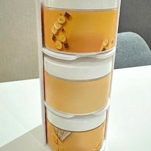 Könnte beinhalten: Ein dreistufiges Behälterset mit weißen Deckeln und transparenten Seiten, gefüllt mit einer gelben Flüssigkeit. Der obere Behälter ist mit Bananenscheiben und Schokoladenquadraten verziert. Der untere Behälter hat Orangenscheiben-Dekorationen.